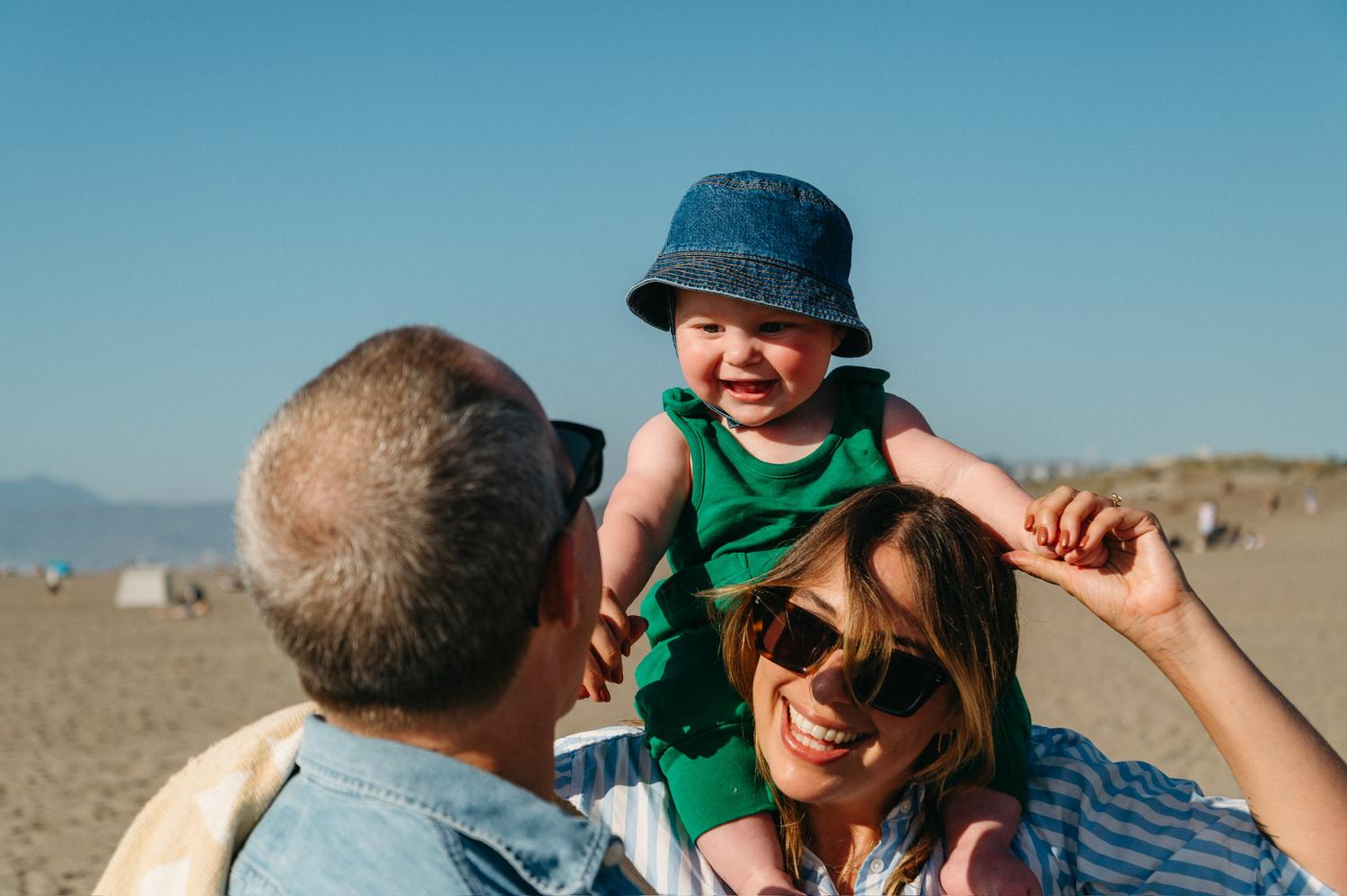 A family at the beach, a parent lifting a smiling baby onto their shoulders